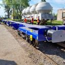 Three quarter trackside view of a new flatbed intermodal carrier wagon going through the final gauge clearance hoop on a sunny day with engineers and trees in background