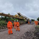 Yellow rail crane arrives on site at Ashington to help install new points for the upgrade project. Engineers in orange suits watch on