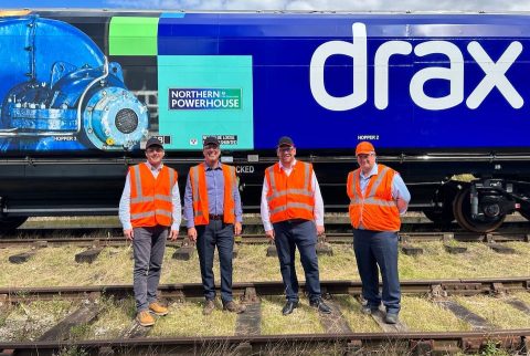 Four gentlemen in orange high visibility vests pose in front of a Drax liveried hopper wagon