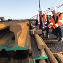 Two men in orange high visibility vests and suits with new rails in a pile in front of them